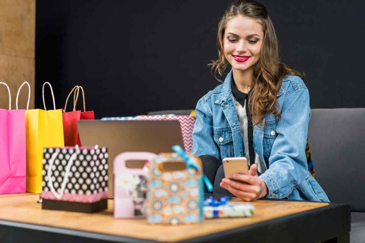 A woman in a denim jacket sitting at a table with shopping bags and a laptop, using her phone.