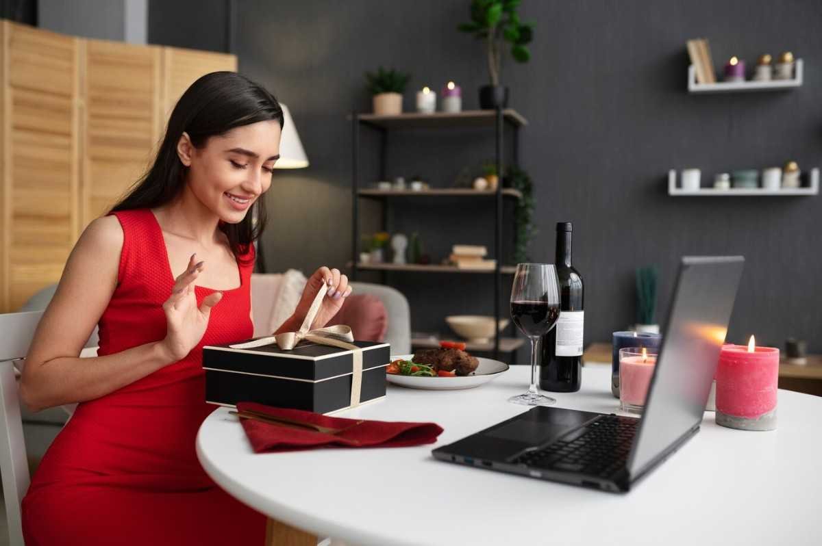 A woman in a red dress smiles while opening a gift box at a dining table. A laptop, wine glasses, and candles are around.